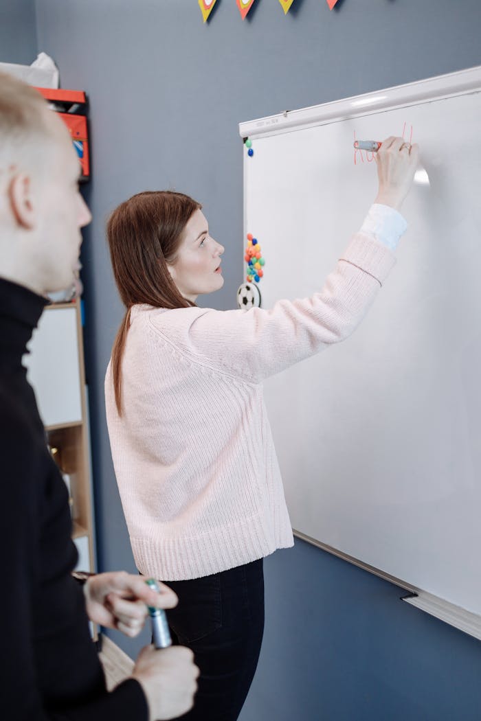 Female teacher writing on a whiteboard, instructing an adult student indoors.